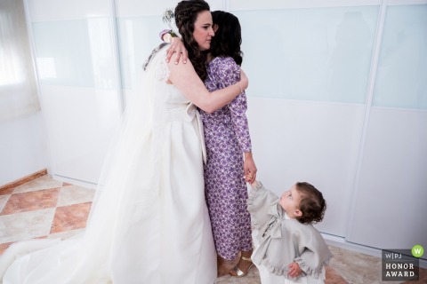 Inside the bride's house in Escañuela, Spain, a young flower girl watches with quiet curiosity as the bride shares a heartfelt and emotional embrace with a beloved family member.