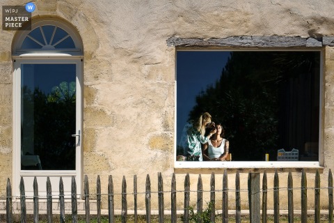 At Château Caillivet in Gironde, France, the photographer masterfully frames the bride indoors through a window during preparations, highlighting anticipation and artistry with a striking outside perspective.