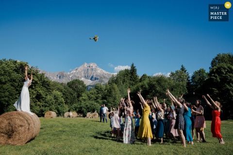 At Gîte de la Marjolière, the photographer captures the bride tossing her bouquet from atop a tall round hay bale, framed against a vivid blue mountain sky, highlighting creative perspective and timing.