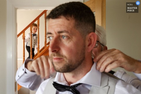 At the groom’s house in Ardara, County Donegal, the photographer captures a poignant scene as the groom’s son watches from the stairs while his grandfather assists the groom, highlighting generational bonds and storytelling skill.