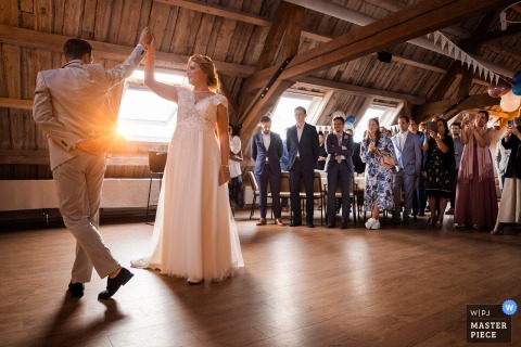 At De Paradijshoeve in Ridderkerk, the photographer captures the bride and groom’s first dance bathed in golden sun rays through a window, surrounded by watching guests, demonstrating masterful use of light and timing.