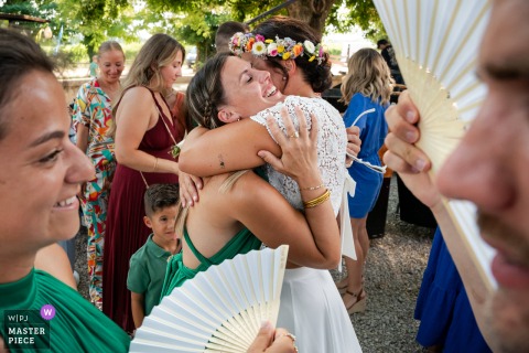 France's Joyful Bride Hugs Guest: A Composition of Friendship In Mauguio, France, the photographer artistically captures the bride hugging a guest, beautifully framed by others in the foreground, showcasing skillful composition and an authentic celebration of friendship.