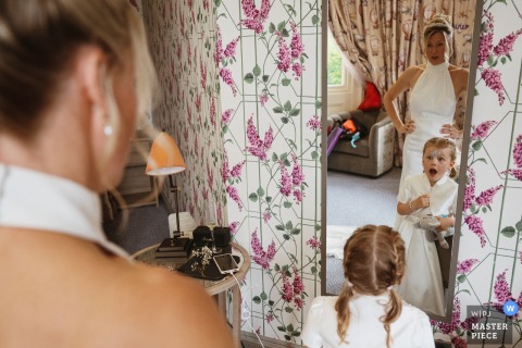 At Pendley Manor Hotel in Tring, UK, the photographer captures the bride’s daughter admiring her reflection in a full-length mirror, with the bride behind her, highlighting touching connection and composition.