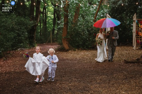At a woodland venue in Herefordshire, UK, the photographer captures joyful children leading the bride and her escort beneath a rainbow umbrella and tree canopy, showcasing expert timing and enchanting storytelling.