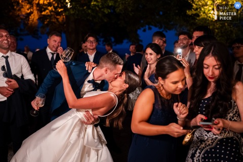 At Baronesse Tacco in San Floriano del Collio, Italy, the photographer captures a layered nighttime scene as the groom surprises the bride with a kiss and casque among guests, demonstrating expert moment-capturing artistry.