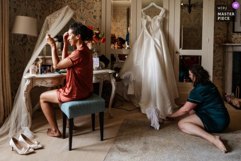 At Hodsock Priory, Nottingham, the photographer captures the bride applying makeup while her bridesmaid tends to the dress, expertly documenting a layered, intimate pre-wedding moment with attention to meaningful details.