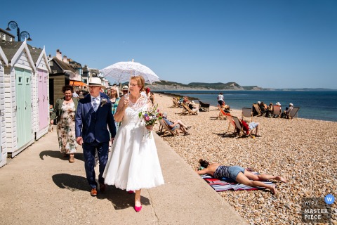 In Lyme Regis, England, a newlywed couple strolls past a sunbather on the beach, the bride holding a white parasol. The photographer’s creative framing contrasts wedding elegance with seaside relaxation.