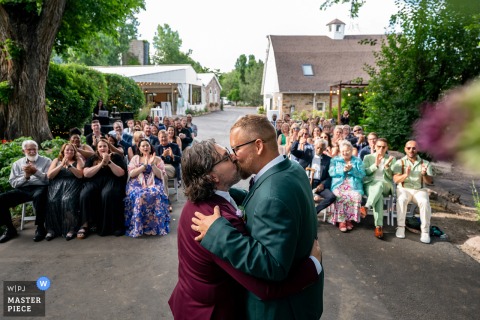 At Tapestry House by Wedgewood Weddings, the couple shares their first kiss, framed by cheering guests. The photographer’s creative use of composition enhances the excitement of the moment.