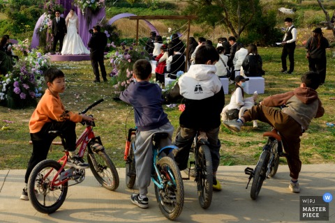 At the ceremony location in Zhanjiang, Guangdong, an outdoor wedding unfolds, bathed in the warm glow of love and celebration as guests and kids on bikes gather under open skies.