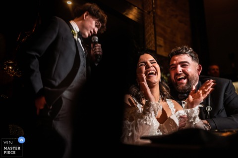 At Ancaster Mill in Ontario, Canada, the couple laughs and claps in a dimly lit room as they react to their son's heartfelt speech during the wedding celebration.