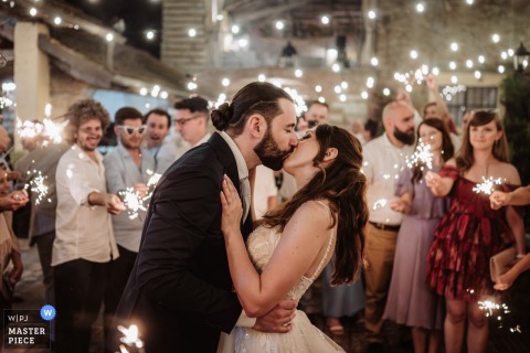 At Cascina Montana in Langhe, Piedmont, the bride and groom share a kiss surrounded by friends holding sparklers under glowing strings of lights during the wedding celebration.