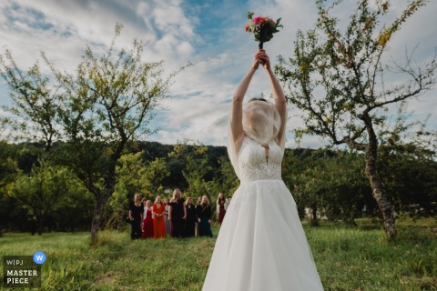 In Meuse at a park during the cocktail hour, the bride prepares to toss her bouquet when the wind blows her stole over her face, creating a lighthearted scene. 