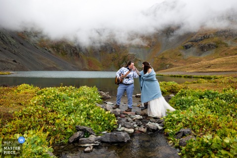   At Clear Lake in Silverton, Colorado, the couple pauses for hand kisses as one helps the other over the rocky creek crossing, highlighting tenderness and connection during their wedding celebration.
