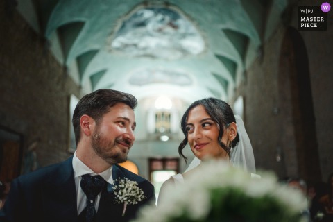 At Badia a Coltibuono in Tuscany, the bride and groom sit together during the ceremony, exchanging gentle, warm smiles in a moment of quiet connection.