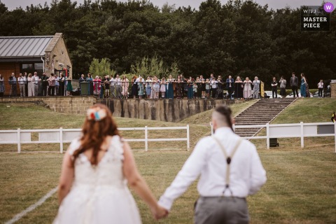 At Harrogate Rugby Club, guests eagerly await the bride and groom’s arrival during an outdoor ceremony, seen from behind in soft focus in the foreground.