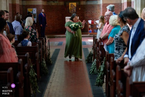 At The Rea Church in Birmingham, a bridesmaid greets a guest with a smile and a wave as she walks down the aisle alone during the wedding ceremony.