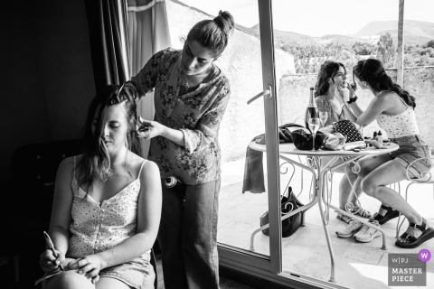 In Valence, France, two women prepare for the wedding, each having hair and makeup done simultaneously by helpers—one indoors and one outside, highlighting the busy pre-ceremony atmosphere.