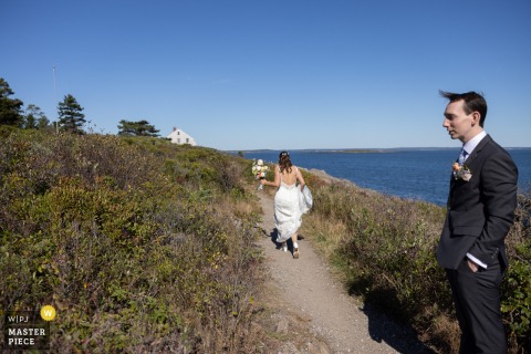 At Giant's Stairs in Harpswell, Maine, the bride walks ahead along the rugged coastline while the groom lingers behind, thoughtfully pausing on their wedding day.