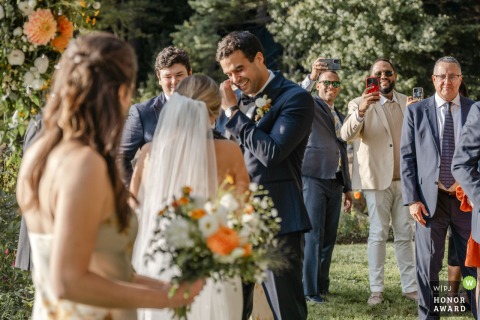 At Migis Lodge in Maine, the groom wipes away tears while eager guests capture photos during the wedding ceremony, preserving heartfelt emotions in every shot.