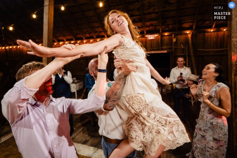 At a wedding in Corvallis, Montana, the bride is held aloft by two men over the dance floor as guests cheer, adding excitement and energy to the celebration.