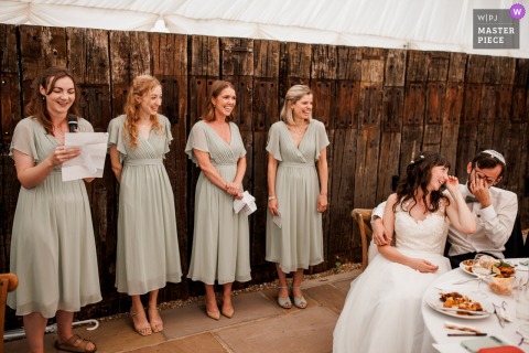 At Burningfold Barn in Godalming, UK, the bride and groom laugh and cover their faces with their hands during the bridesmaids’ speeches at the wedding reception.