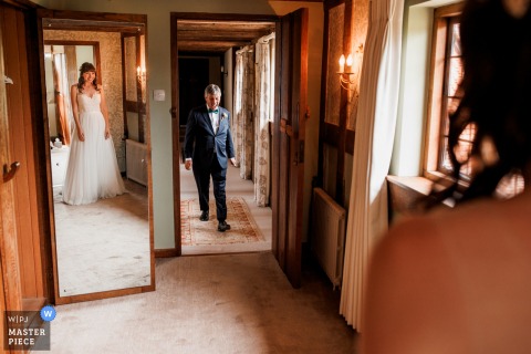 At Burningfold Barn in Godalming, UK, the father of the bride walks down the hall and sees his daughter for the first time, her reflection visible in a mirror nearby.