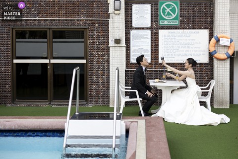 At Asia Hotel in Bangkok, the bride and groom enjoy a quiet break, sitting poolside at a table and facing each other during their wedding celebration.