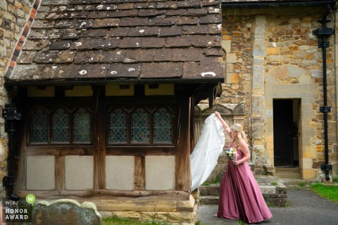Outside the St Nicholas Church in Worth, West Sussex, a dramatic and elegant scene is created by the bride’s long wedding train trailing gracefully held by bridesmaid.