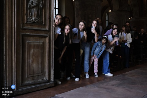 At Chiaravalle Abbey in Milano, Italy, a row of girls waits inside the church for their teacher, who is about to get married, glancing curiously over each other.