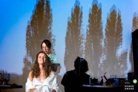In a sunlit room at Château de Beauséjour, the bride has her hair styled while the park outside is reflected in the window, creating a dreamy, ethereal atmosphere.
