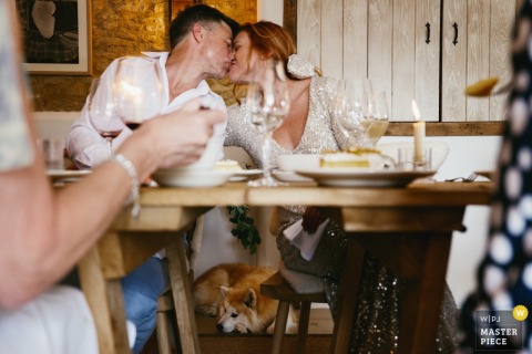 The couple shares a loving kiss during their intimate wedding lunch at The Bell in Charlbury, Cotswolds, while their faithful dog naps contentedly on the floor between them.