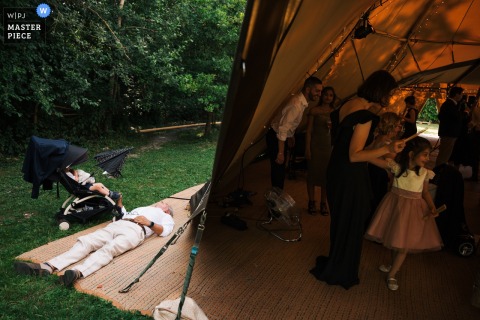 Outside the tipi at Cress Lake wedding venue, Hertfordshire, the bride's father gently snoozes while keeping watch over his young grandson sleeping soundly in a pram.