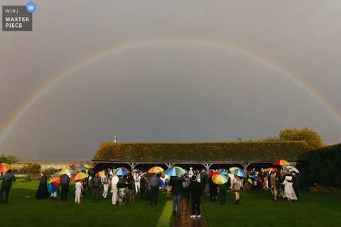 At Elmley Nature Reserve, Isle of Sheppey, guests enjoy a rainy drinks reception, finding shelter under colorful umbrellas beneath a spectacular, wide double rainbow.