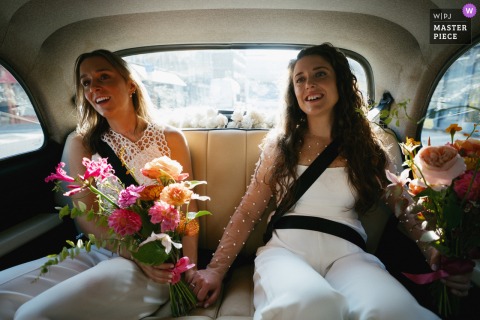 Two brides travel to their Islington Town Hall wedding ceremony in a London taxi, beautifully illuminated by the warm, late afternoon sunshine streaming through the windows.