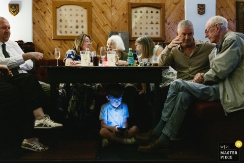 At the MOTH Club evening reception in Hackney, London, family members chat around a table while a young guest is absorbed by his phone's blue light underneath.