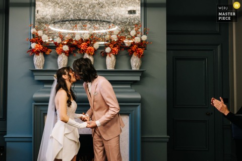 At a London reception venue, the bride and groom share their first kiss indoors before a grand fireplace, marking a romantic highlight of their wedding day.