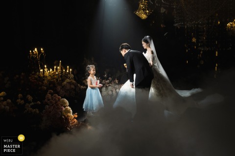 At Xijiao State Guest Hotel in Shanghai, China, under a spotlight in the dark setting, the newlyweds bend down to warmly greet the flower girl bringing the rings—a romantic and touching moment.