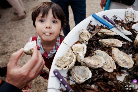 In Kerlouan, France, a little girl tries an oyster for the first time, her expression full of curiosity during the wedding celebration.