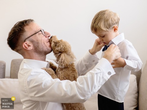 At the groom's home in Lombardia, Italy, a dad fixes his son's shirt while their small dog licks his face, adding a humorous, heartwarming moment to the wedding preparations.