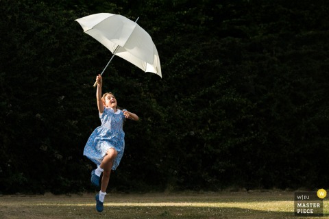 At a family farm reception venue in Northamptonshire, East Midlands, a flower girl joyfully plays with an umbrella against a dark background, in a minimalist frame full of childhood charm.