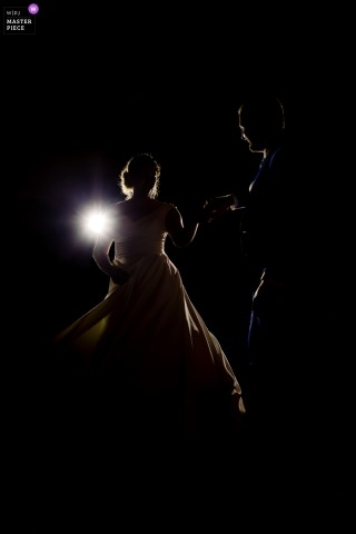 In Saint Alban Leysse, Savoie, France, the bride and groom share their first dance, backlit and rimlit against a dark background in a dramatic vertical shot.