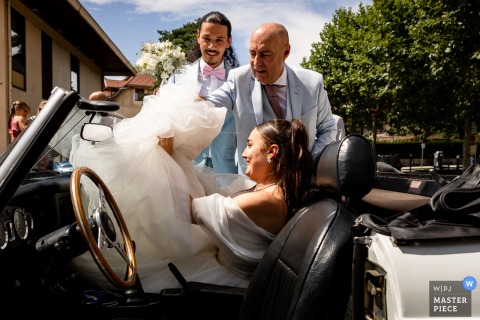 At the city hall of Savoie, France, the bride smiles in the sunshine as her fluffy dress struggles to fit into a vintage convertible, adding a playful touch to the wedding day.