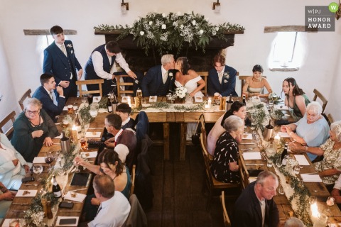 In Cruck Barn at the Craven Arms, Appletreewick, North Yorkshire, the bride and groom share a secret kiss while guests settle in for the wedding breakfast.