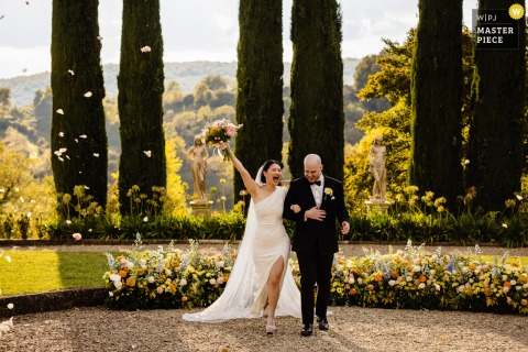 At Villa La Selva in Tuscany, the couple exits their ceremony as petals fly in the sunlight, framed by tall cypress trees in the background.