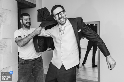 During a wedding in Poitou-Charentes, a black-and-white shot shows the groom receiving help with his jacket during a tricky preparation moment, highlighting the candid challenges and teamwork before the ceremony.
