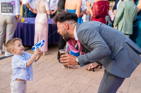 At Salones Victoria in Torreperogil, Jaén, Spain, a mischievous boy playfully sprays water in a man's face during the wedding, a candid and humorous scene amid the festivities.