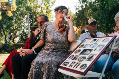 At Danwalt Gardens, the groom’s mother sits tearfully beside a picture board honoring the groom’s late father during the wedding ceremony, capturing a poignant moment of remembrance and emotion.