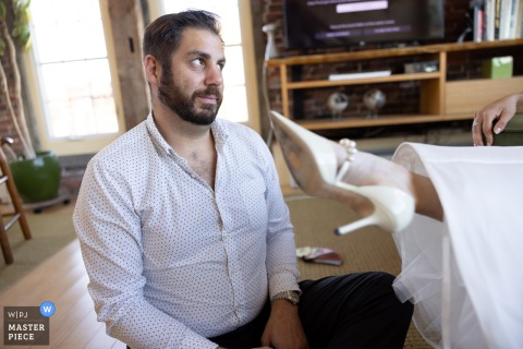 At an Airbnb in Portland, Maine, the groom’s brother thoughtfully helps the bride with her shoes, capturing a warm and supportive moment of family connection during wedding preparations.