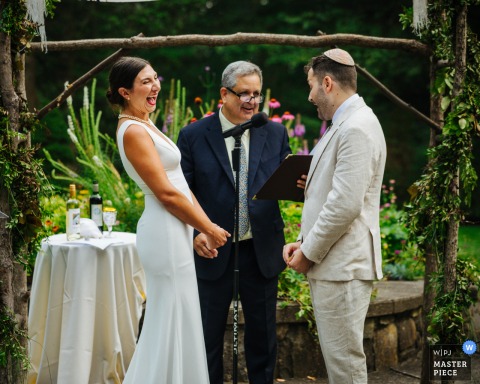 At Arrow Park, NY, the couple bursts into laughter during their vows at the altar, surrounded by a lush garden setting and arbor, capturing joy and genuine connection in the middle of their ceremony.