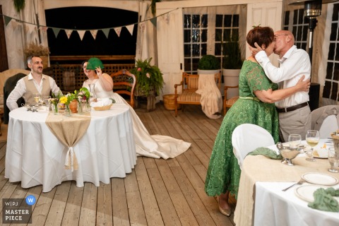 At La Maison d'Évelyne in Quebec, Canada, the bride’s parents stand and share a kiss during dinner while the newlyweds playfully cover their eyes, capturing a joyful and lighthearted family moment at the reception.